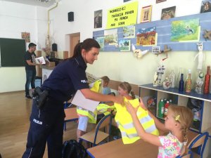 Na zdjęciu policjantka podchodzi do uczniów siedzących w ławkach i wręcza kamizelki odblaskowe.