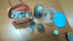 Na stole torba z popakowanymi w torebki foliowe narkotykami, dwa słoiki z marihuaną, dwie nakrętki do słoików, waga elektroniczna.
