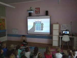 Dzieci oglądają film edukacyjny pod tytułem Miś Poli na ekranie widać telefon alramowy 112