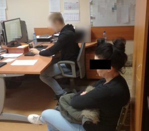 Policjant siedzi przy biurku i pisze na komputerze, zatrzymana 48-letnia kobieta siedzi obok na krześle.
