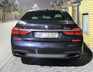 Na zdjęciu samochód marki BMW koloru czarnego zaparkowany przy ogrodzeniu. Zdjęcie przedstawia tył samochodu.