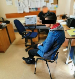 Na zdjęciu, w pomieszczeniu na krześle siedzi zatrzymany mężczyzna, obok widać policjanta siedzącego przy biurku. Policjant przesłuchuje mężczyznę.