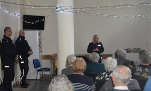 Na zdjęciu osoby starsze zgromadzone w świetlicy klubu seniora, na pierwszym planie widać policjantów z Bielan, którzy opowiadają seniorom o zasadach bezpieczeństwa.