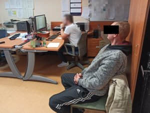Na zdjęciu, w pomieszczeniu komendy na krześle siedzi zatrzymany mężczyzna, na dalszym planie widać policjanta siedzącego przy komputerze i przesłuchującego zatrzymanego.
