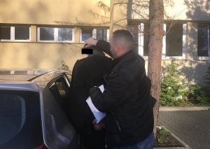 Na zdjęciu parking policyjny, policjant pilnuje zatrzymanego, pomaga mężczyźnie do wsiąść do radiowozu nieoznakowanego.
