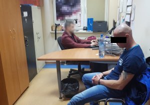 Na zdjęciu widać pomieszczenie w którym na krześle siedzi zatrzymany mężczyzna, na dalszym planie przy komputerze siedzi policjant, przesłuchujący zatrzymanego.