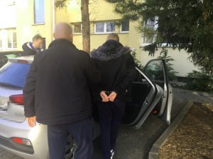 Na zdjęciu na policyjnym parkingu policjant prowadzi zakutego w kajdanki zatrzymanego mężczyznę do radiowozu nieoznakowanego. Z drugiej strony auta znajduje się drugi policjant. Policjanci nie są umundurowani.