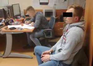 Na zdjęciu zatrzymany mężczyzna siedzi na krześle, obok przy biurku policjant przesłuchujący mężczyznę, piszący przy komputerze.