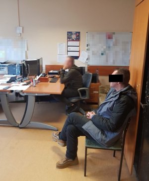 Na zdjęciu w jednym z policyjnych pomieszczeń na krześle siedzi zatrzymany mężczyzna. Na drugim planie przy biurku siedzi przesłuchujący go policjant.