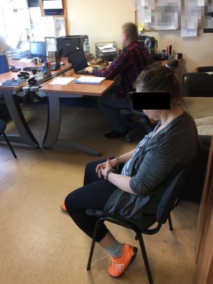 Na zdjęciu w pomieszczeniu komendy na krześle siedzi zatrzymana kobieta, na dalaszym planie policjant siedzący przy biurku.