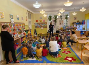 Na zdjęciu w sali przedszkola na dywanie siedzą przedszkolaki, obok na krzesełkach nauczycielki. Na dalszym planie stoi policjantka prezentuje dzieciom elementy wyposażenia policjanta do służby.