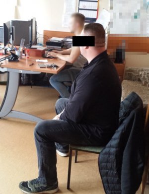 Na zdjęciu jedno z pomieszczeń w komendzie w którym na krześle siedzi zatrzymany mężczyzna, na dalszym planie, przy biurku siedzi policjant i pisze na komputerze.