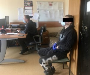 W pomieszczeniu na pierwszym planie znajduje się zatrzymany mężczyzna, siedzi na krześle, na ręce założone ma kajdanki. Na drugim planie widać policjanta siedzącego na krześle przy biurku. Policjant zapisuje na komputerze zeznania zatrzymanego.