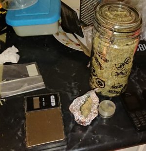 Na zdjęciu widoczny szklany słój z zawartością marihuany, waga elektroniczna oraz pudełko i zawiniątko z marihuaną.