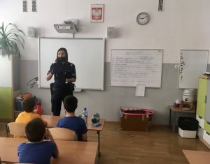 Policjantka w sali lekcyjnej podczas akcji Lato w mieście opowiada o zasadach bezpieczeństwa. Funkcjonariuszka ma założoną maseczkę oraz rękawiczki.