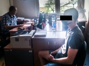 Na zdjęciu w pokoju policjant przesłuchuje zatrzymanego mężczyznę. Zatrzymany widoczny jest na pierwszym planie, siedzi na krześle. Dalej widać siedzącego przy biurku z komputerem policjanta.