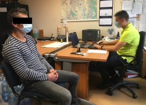 Na zdjęciu na pierwszym planie na krześle siedzi zatrzymany mężczyzna. Na drugim planie widać policjanta siedzącego przy biurku.