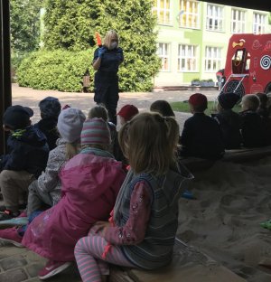 Na zdjęciu na pierwszym planie siedzące na ławeczkach przy piaskownicy przedszkolaki zwrócone w stronę policjantki. Policjantka prezentuje najmłodszym policyjną latarkę.