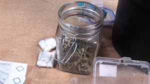 Na pierwszym planie szklany słoik z zawartością marihuany, w tle częściowo widoczne banknoty, woreczki foliowe z kokainą oraz plastikowe pudełko.