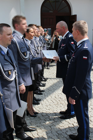 Policjanci stoją w szeregu. Policjant wręcza policjantce dokument.