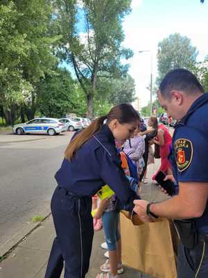 Policjantka sięga ręka do torby, która trzyma strażnik miejski. W tle grupa dzieci.
