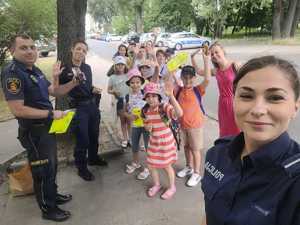 Strażnik miejski trzyma w rękach kamizelkę odblaskową i telefon komórkowy. Policjantki pozują do zdjęcia z grupą dzieci i opiekunką.