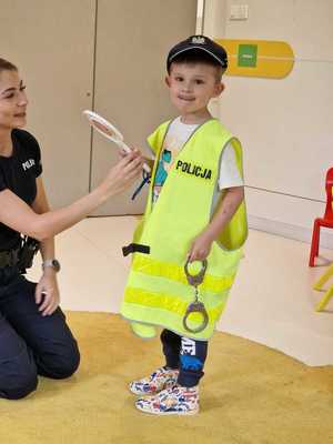 Policjantka i chłopiec w kamizelce odblaskowej. Chłopiec w jednym reku trzyma kajdanki, w drugim tarczę do zatrzymywania pojazdów.