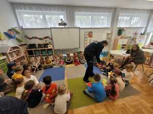 Policjant rozdaje książeczki siedzącym dzieciom.