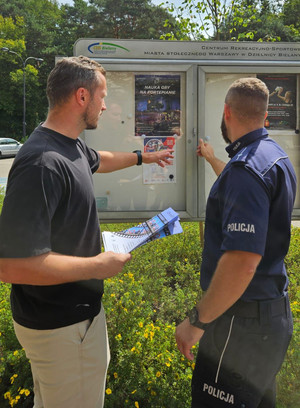 Policjant z pracownikiem stoją przed tablicą ogłoszeniową.